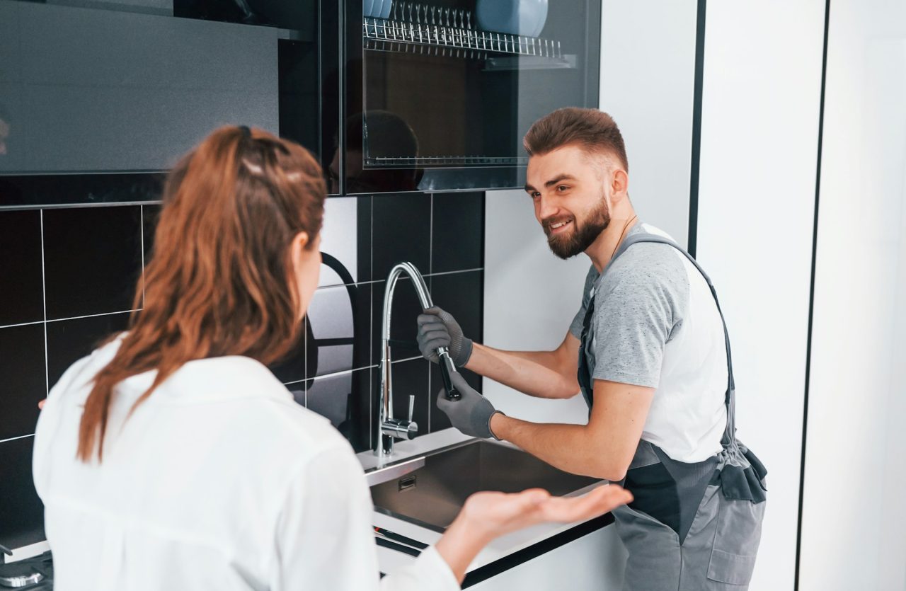 young-professional-plumber-in-grey-uniform-shows-results-of-work-for-housewife-on-the-kitchen